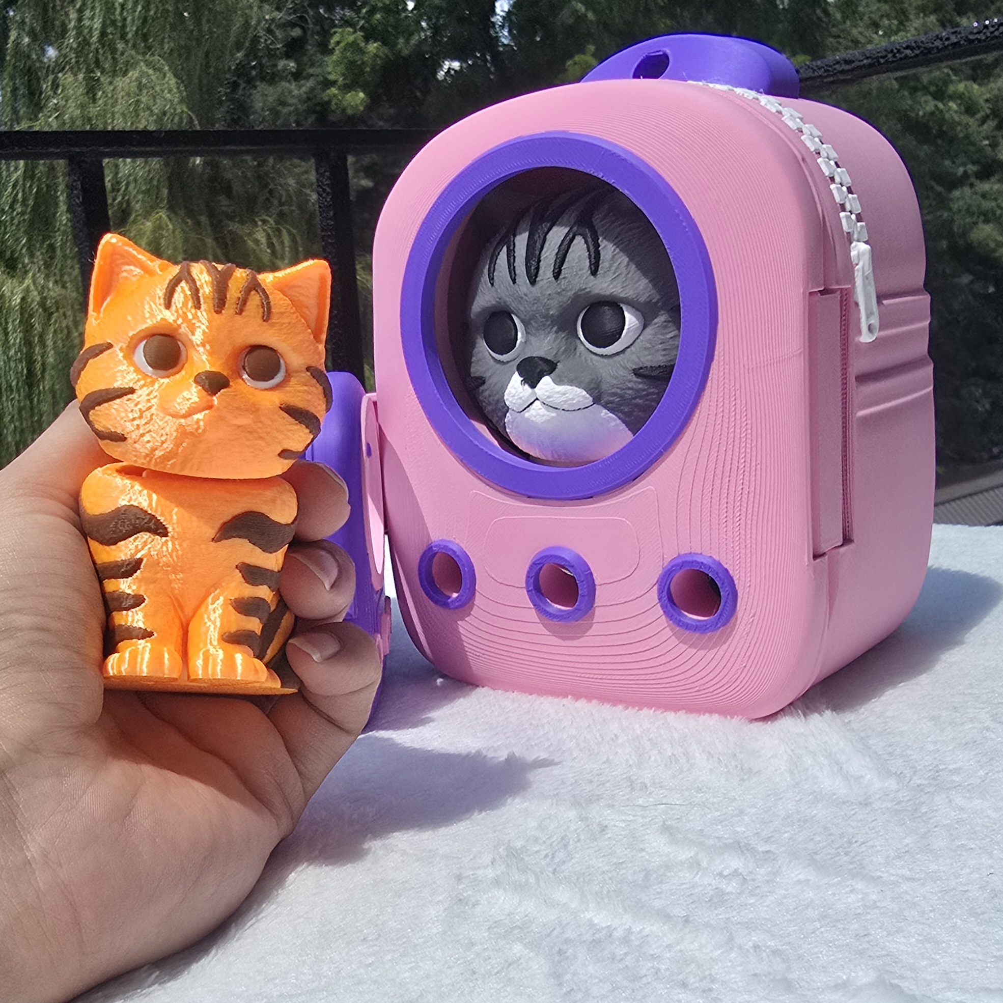A 3 inch orange tabby cat print held in a hand beside a pink backpack with a gray cat in the window