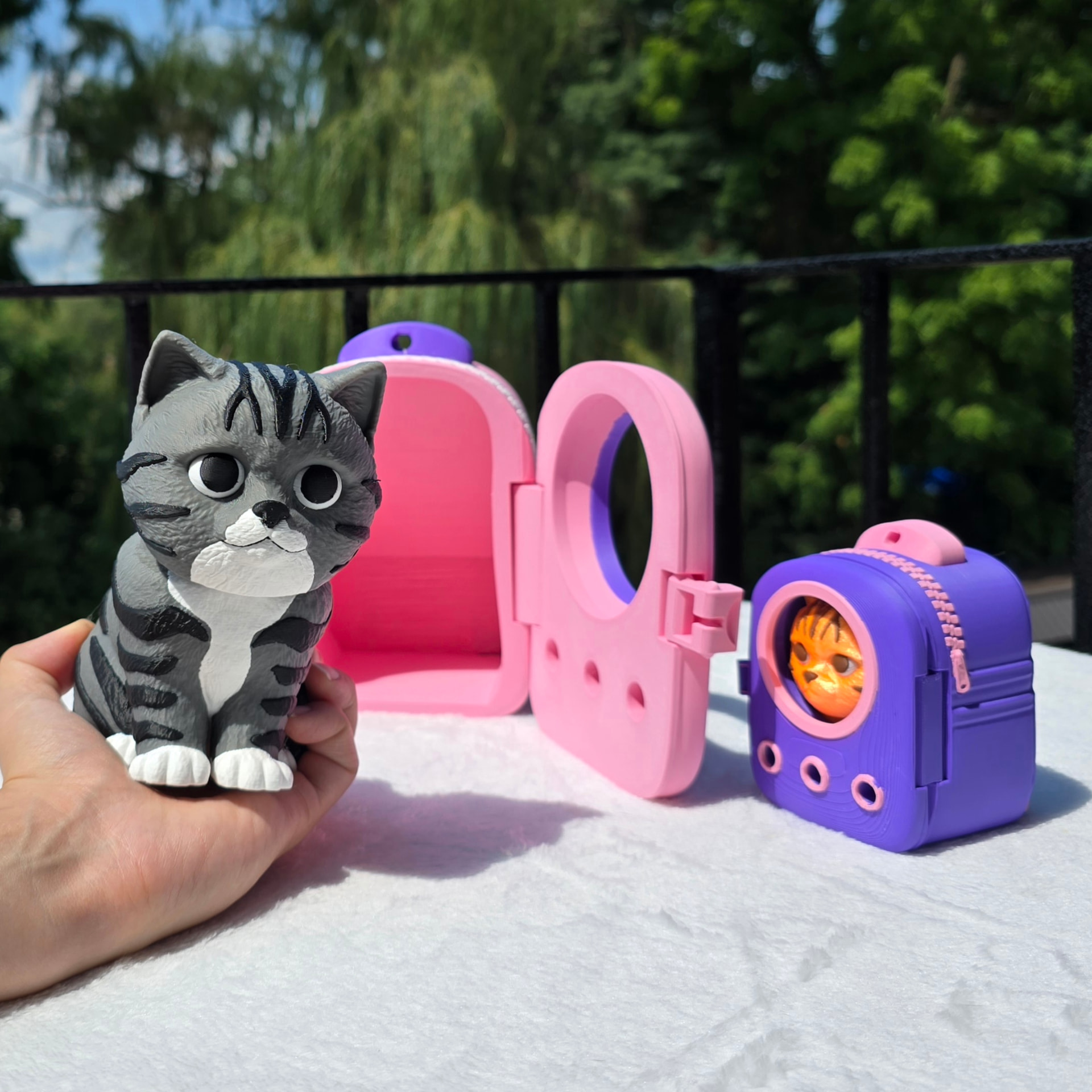 A 6 inch gray tabby cat print held in a hand beside an open pink backpack and a purple backpack with an orange cat inside